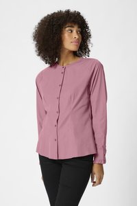 Chemise à manches longues boutonnée en tissu rose clair, avec un col rond et des poignets à volants, portée avec un pantalon noir ajusté.