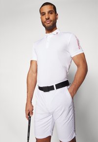Vit pikétröja med krage och tre knappar, med röd logotyp på ärmen. Matchad med vita shorts och ett svart bälte, hållande en golfklubba.