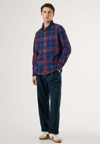 Camisa de cuadros azul marino con un patrón de rejilla rojo y blanco, abotonada, de manga larga. Combinada con pantalones azules oscuros y zapatillas blancas.