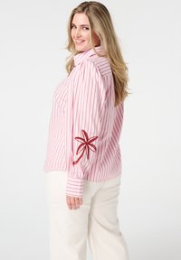 Roze en wit gestreepte blouse met lange mouwen, voorzien van een geborduurd bloemdetail op de linkerarm en met knoopsluiting bij de manchetten.