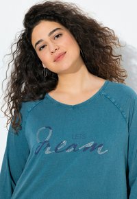 Tyrkysová dlouhá rukávová košile s výstřihem do V a raglánovými rukávy, se třpytivým nápisem: "LETS DREAM" v modré a stříbrné barvě napříč hrudníkem.