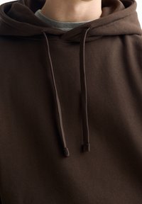 Felpa marrone con una texture morbida, con cordini e cappuccio. Indossata sopra una maglietta grigia, con una vestibilità comoda e un design semplice.