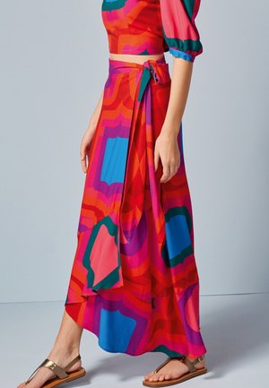 Vrouw draagt een kleurrijke wikkelrok met geometrische print en een gestrikte taille en middellange pofmouwen, gecombineerd met bruine platte sandalen.