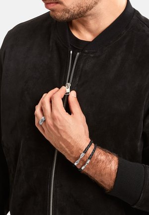 Mann trägt eine schwarze Wildlederjacke, die teilweise zugezippt ist, zeigt geflochtene schwarze Lederarmbänder und einen silbernen Ring an der linken Hand.