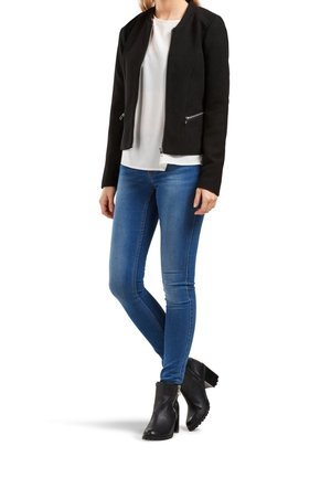 Kvinde iført en sort jakke med lynlås, hvid bluse, blå tætsiddende jeans og sorte ankelstøvler, stående med det ene ben krydset foran det andet.