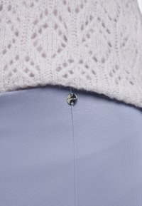 Hellgrauer Strickpullover mit einem Spitzenmuster über einem glatten Lavendelfarbenen Rock, der mit einem metallischen Knopfdetail am Bund versehen ist.