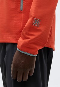 Σκούρο δέρμα σε χέρι που φοράει πορτοκαλί μπλούζα με μακριά μανίκια και γκρι μανσέτα, με κεντημένο γράμμα "R" στο μανίκι, συνδυασμένη με μαύρο παντελόνι.