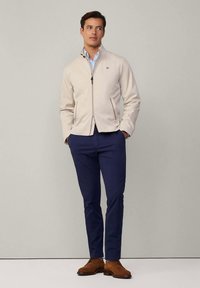 Ljusbeige zip-up-jacka med ståkrage, två sidofickor och broderad logotyp. Kombinerad med marinblå byxor och bruna snörskor.