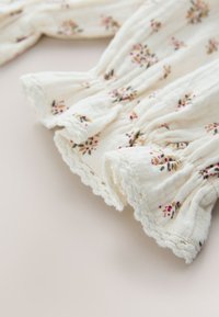 Tela blanca con pequeños estampados florales en rosa y amarillo, bordes elásticos fruncidos y ribete de encaje con borde ondulado en mangas o puños.
