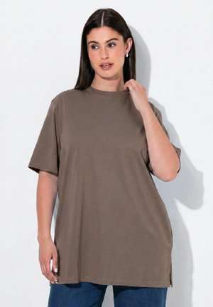 Femme aux longs cheveux foncés portant une chemise marron oversize à manches courtes et un jean bleu, debout sur un fond blanc.