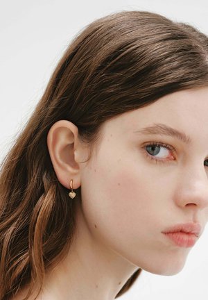 Joven mujer con piel clara y cabello castaño, que lleva pendientes colgantes pequeños de oro en forma de corazón, mirando hacia un lado con un fondo liso.