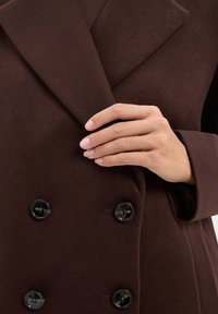 Dunkelbrauner doppelt geknöpfter Wollmantel mit großen schwarzen Knöpfen, breiten Revers und einem strukturierten Stoff. Eine Hand ruht auf der Vorderseite des Mantels.