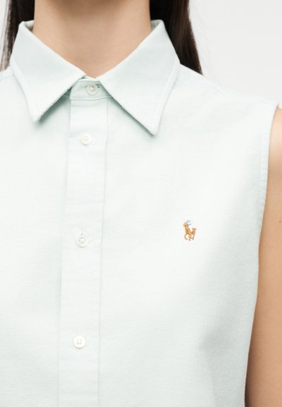 Gros plan sur une personne portant une chemise sans manches bleu clair à boutons, avec un petit logo brodé d'un poney marron sur la poitrine.