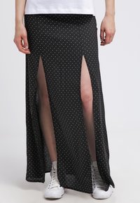 Černá maxi sukně s puntíky a vysokými rozparky, vyrobená z lehkého materiálu, v kombinaci s bílými teniskami. Má přiléhavý pas a splývavý design.