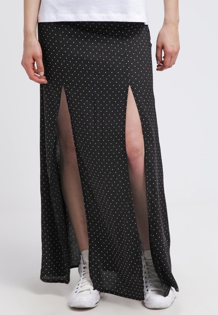 Černá maxi sukně s puntíky a vysokými rozparky, vyrobená z lehkého materiálu, v kombinaci s bílými teniskami. Má přiléhavý pas a splývavý design.