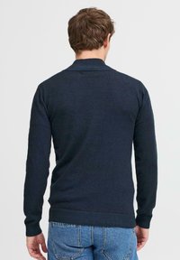 Sötét tengerészkék kötött pulóver bordázott gallérral és mandzsettával, hosszú ujjakkal és testhez simuló fazonnal, farmerrövidnadrággal párosítva.