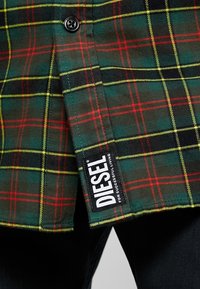 Grön, röd och gul rutigt tyg med svart knapp och svart etikett med texten "DIESEL For Successful Living" på fållen av plagget.
