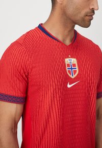 Rød Nike fodboldtrøje med tekstureret design, V-hals, Norges nationalmandskabs emblem og blå accenter på ærmerne.