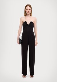 Combinaison noire avec un décolleté en V plongeant et des détails froncés au niveau de la poitrine. Design à jambes larges et longues. Le mannequin tient une petite pochette noire.