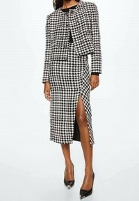 Completo houndstooth bianco e nero con giacca corta e gonna al di sotto del ginocchio con spacco laterale, abbinato a décolleté neri a punta.