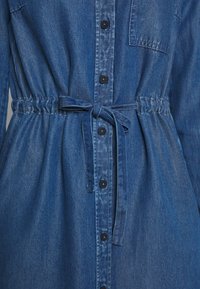 Denimklänning med framknappar, elastisk midja, knytbälte och bröstficka, som visar en texturerad blå tyg och sömnadsdetaljer.