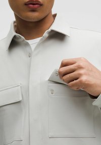 Hele halli nööbitava särk tekstuurse kangaga, millel on kaks eesmist taskut, terav krae ja nööbiga kinnitamine rindetaskus.