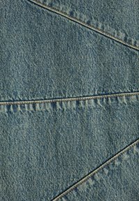 Denim tkanina plave boje, s teksturiranom površinom, vidljivim šavovima i dijagonalnim šavnim uzorkom koji prolazi kroz tkaninu.