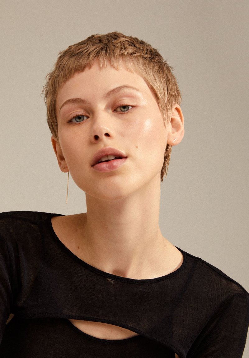 Haut transparent noir avec une découpe arrondie au niveau du col, entourant le visage d'une personne aux cheveux courts et ébouriffés, portant de simples boucles d'oreilles de type clou.