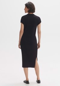 Abito midi nero a coste con maniche corte, fessure laterali e silhouette aderente. Modella vista di spalle, design minimalista.