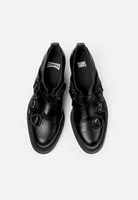 Zapatos de cuero negro sin cordones con cintas elásticas anchas y acentos de hardware redondeados. Suelas de goma planas, diseño minimalista y acabado suave.