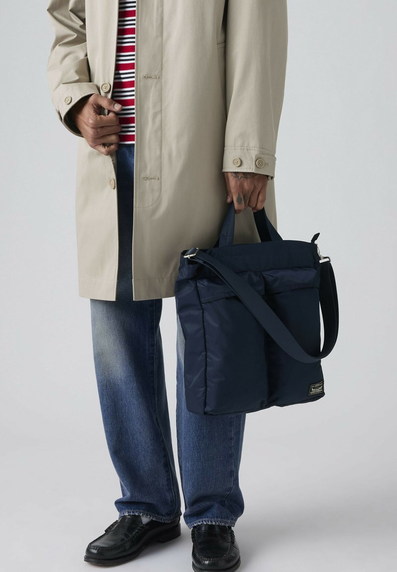 Persona con gabardina beige, camisa de rayas, jeans azules y zapatos negros, sosteniendo un bolso tote azul marino con asas y correa para el hombro.