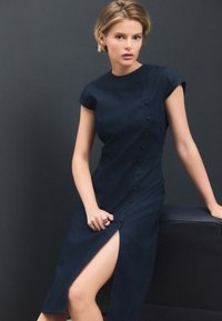 Robe en denim bleu marine avec un col montant, des manches courtes et une fente latérale. Présente une rangée de boutons sur le devant et une silhouette ajustée.