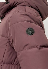 Burgunderfarbene Steppjacke mit gestepptem Design, ausgestattet mit einem schwarzen runden Logo-Patch am Ärmel und einer strukturierten, glatten Stoffoberfläche.
