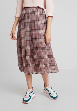 Mujer con falda midi plisada rosa con patrón de cuadros, blusa rosa claro y zapatillas deportivas blancas, turquesas y rosas sobre un fondo gris.