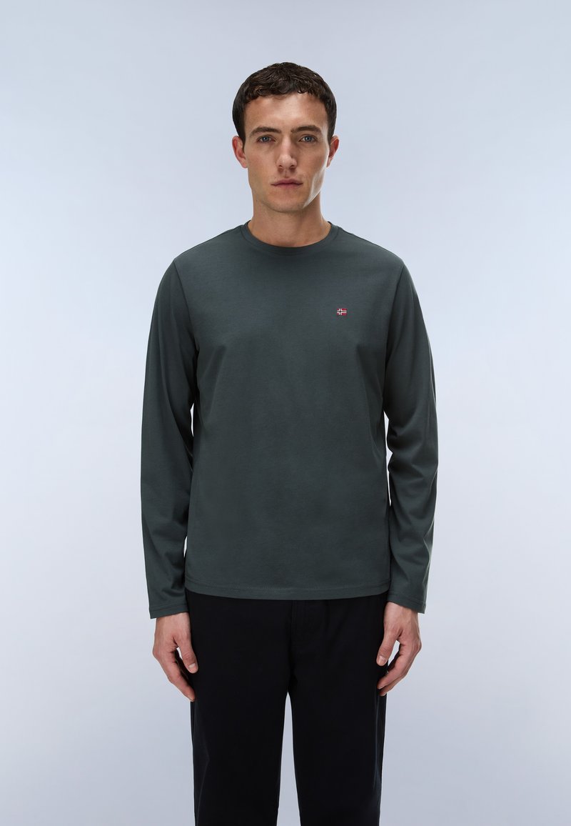 Jonge man met kort donker haar, wearing een donkergroen longsleeve met een klein vlaglogo op de borst en zwarte broek.