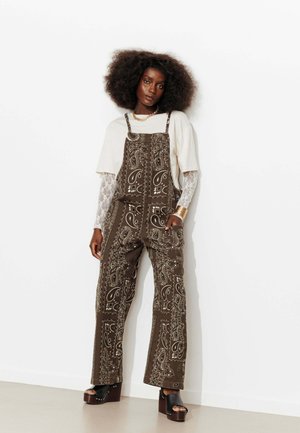 Femme aux cheveux bouclés et volumineux portant des manches en dentelle blanche sous un top blanc ample, une salopette marron à motif cachemire et des sandales à plateforme.