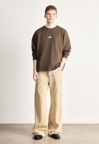Felpa marrone con logo, abbinata a pantaloni beige larghi e stivali neri. Tessuto liscio, vestibilità comoda e design minimalista.
