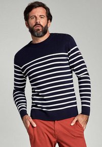 Pullover a righe blu navy e bianche con scollo rotondo, maniche lunghe, dettagli con bottoni sulle spalle, indossato con pantaloni rossi.