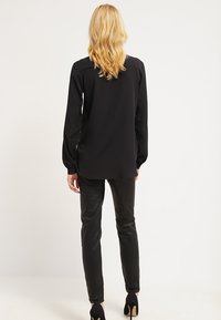 Svart långärmad blus med avslappnad passform, slät tyg och knäppningar vid ärmsluten, kombinerad med glänsande svarta slim-fit byxor och högklackade skor.