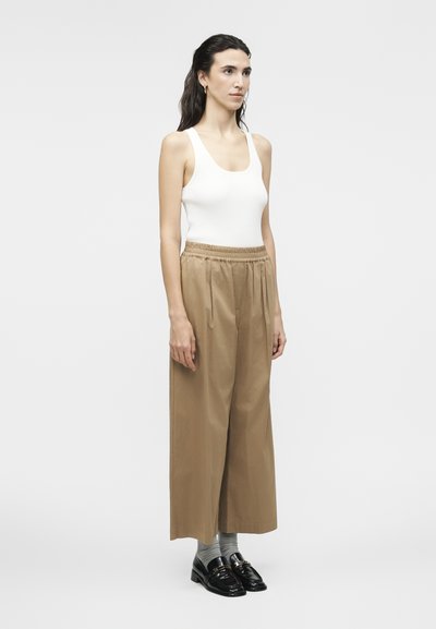 Γυναίκα όρθια φορώντας λευκό αμάνικο τοπ, μπεζ παντελόνια με φαρδιά μπατζάκια, γκρίζες κάλτσες και μαύρα loafers σε απλό φωτεινό φόντο.