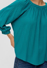 Bluza w kolorze niebiesko-zielonym z teksturowanym materiałem, w pionowe paski, z zebranym dekoltem i bufkami. Dół delikatnie zaokrąglony. Noszona z ciemnymi dżinsami.