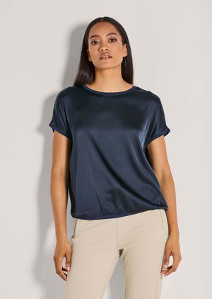 Frau mit glattem dunklem Haar, die eine dunkelblaue kurzärmelige Satinbluse und hellbeige eng anliegende Hose trägt, stehend vor einem einfarbigen Hintergrund.