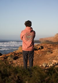 Felpa rosa con un design grafico circolare sulla schiena, abbinata a pantaloni scuri; in piedi su un terreno roccioso vicino alla costa.