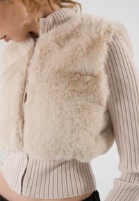 Vest scurt din blană artificială bej, cu guler și tiv din tricot cu model în relief. Suprafață texturată cu o strălucire subtilă, închidere cu fermoar pe toată lungimea și design ajustat.