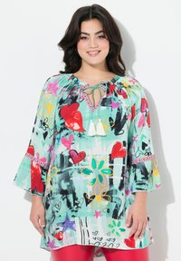 Tunique colorée avec un design fluide, présentant un fond vert menthe, des motifs floraux et en forme de cœur, ainsi que des accents à franges au niveau du col.