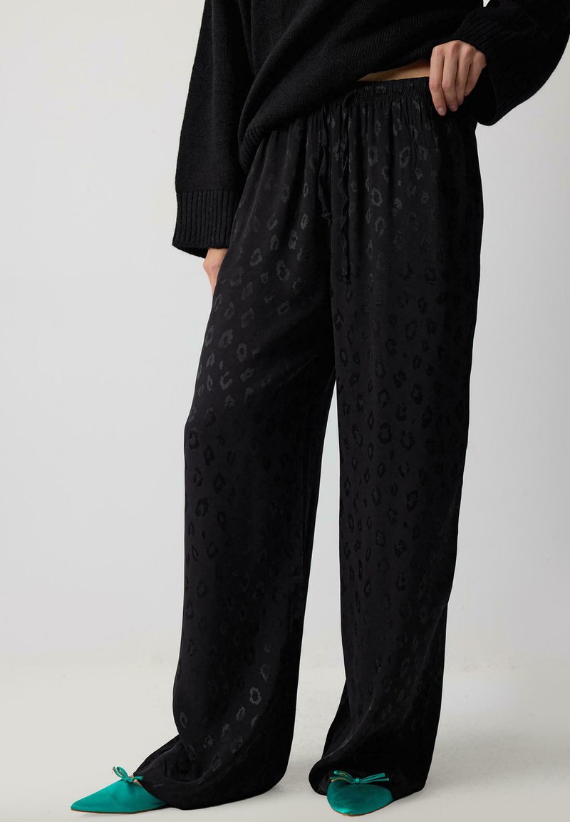 Black leopard print pants with a relaxed fit, elastic waistband, and a soft, textured fabric. Paired with pointed teal shoes.