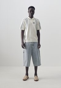 Polo shirt in white knit with dark stripes, paired with light blue denim cargo shorts. Tan slip-on shoes with textured detailing.