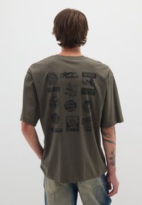 Camiseta de gran tamaño color verde oliva, material de algodón, con logotipos y gráficos impresos en negro en la parte posterior, mangas cortas y escote redondo.