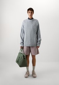 Hellblaues Kabelstrickpullover, lilafarbene Shorts, graue Kniestrümpfe und rosa Sportschuhe mit einem grünen Tragetasche, die grifffreie Henkel und eine Fronttasche hat.