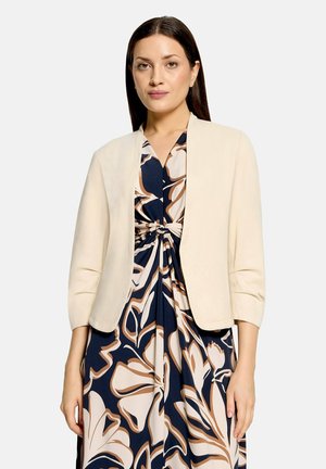 Kvinde med mørkt hår iført en beige kort jakke over en mørkeblå kjole med store beige og brune blomsterprint.